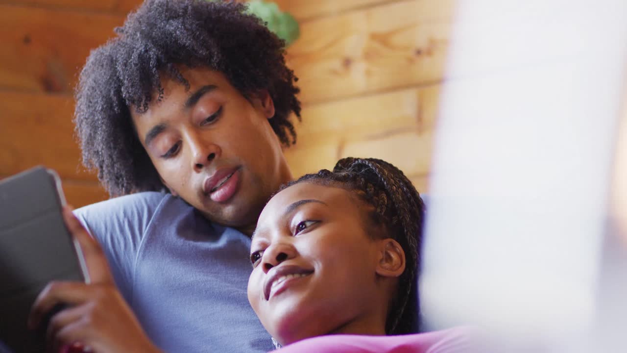Coppia afroamericana felice che usa il tablet, seduta sul divano in una baita di legno, al rallentatore