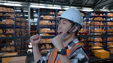 vista lateral de cerca de un ingeniero masculino asiático con casco de seguridad de pie y gritando meta celebrando trabajando en el almacén con estantes llenos de mercancías de entrega