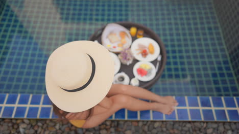 top view on the gorgeous slim girl in straw hat sitting poolside with her legs tucked up beneath one, lunch set up next to the pool