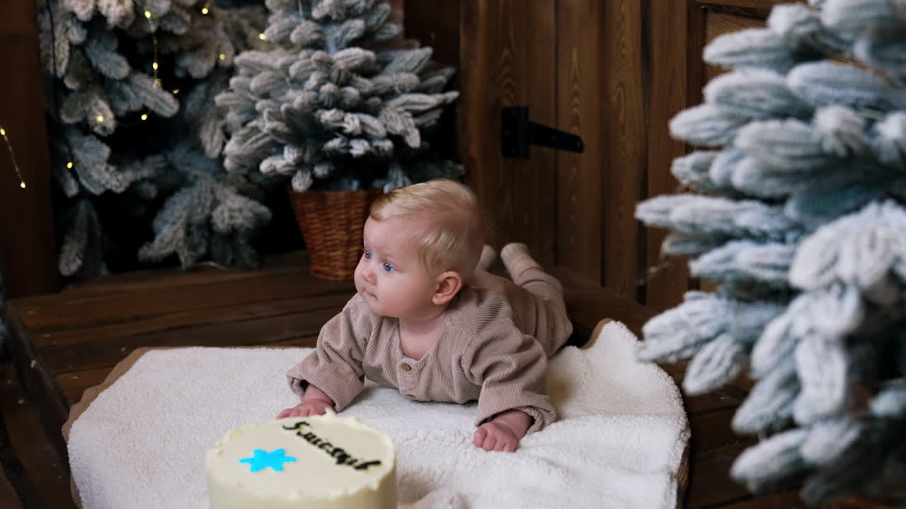 Lovely cute little baby with fair hair lies on the soft plaid. Big cake and Christmas trees around the child.