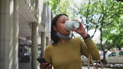 african american woman wearing face mask using smartphone drinking coffee in street