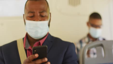 African-american-man-in-city-wearing-face,-using-smartphone-in-bus