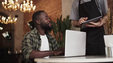 A-black-person-with-a-beard-works-on-a-laptop-in-a-cafe,-a-waiter-approaches-them-and-takes-an-order