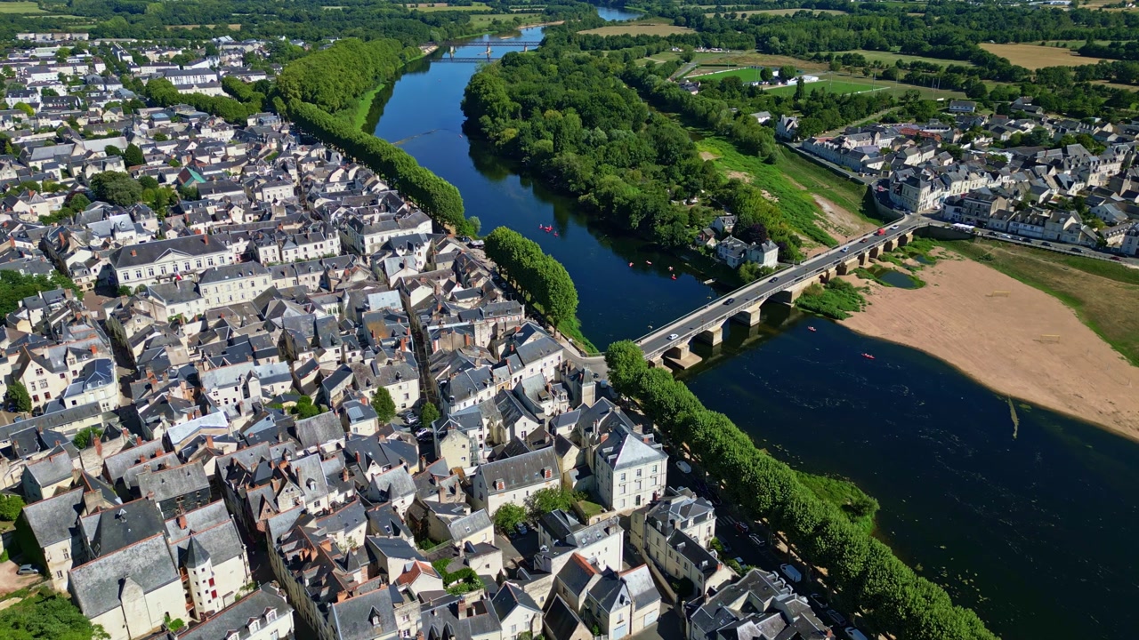 Drone top view of Chinon showing the bridge over the Vienne River, town rooftops, riverside vegetation, and surrounding fields.