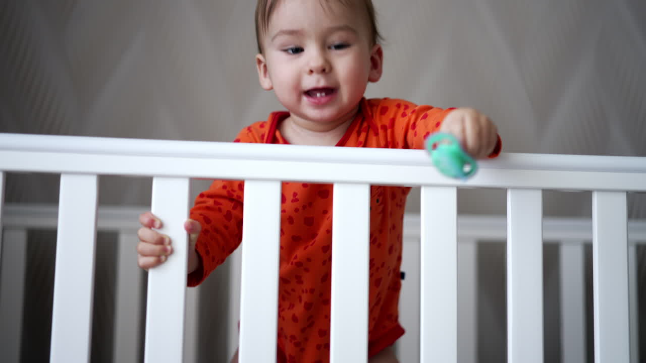 Sweet funny little baby standing in white crib. Smiling one year old toddler throwing his pacifier happily and gets it back.