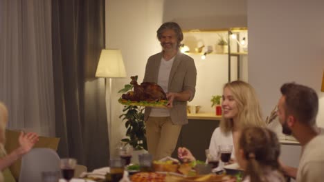 gray haired man brings the cooked turkey to the table where the family is sitting to celebrate thanksgiving