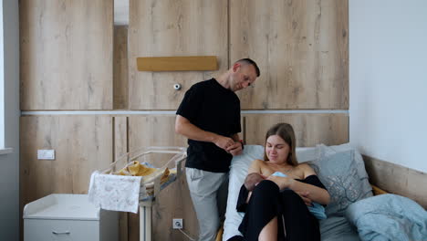 long-haired woman sits in hospital bed breastfeeding her newborn. man stands beside admiring the view from top.