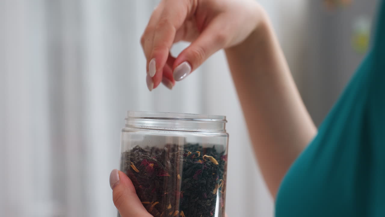 Caucasian Woman Picking Loose Tea Jar Fingertips Dip Into Glass Container Retrieving Dried Leaves, CloseUp Of Tactile Selection And Aroma Discovery, Relaxed Morning Tea Routine In Home Kitchen