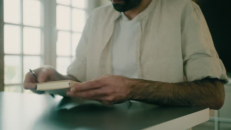 un joven creativo pensativo entra en su sala de estar, se sienta en una mesa junto a una ventana, abre un pequeño cuaderno y comienza a escribir planes, ideas o listas con un bolígrafo