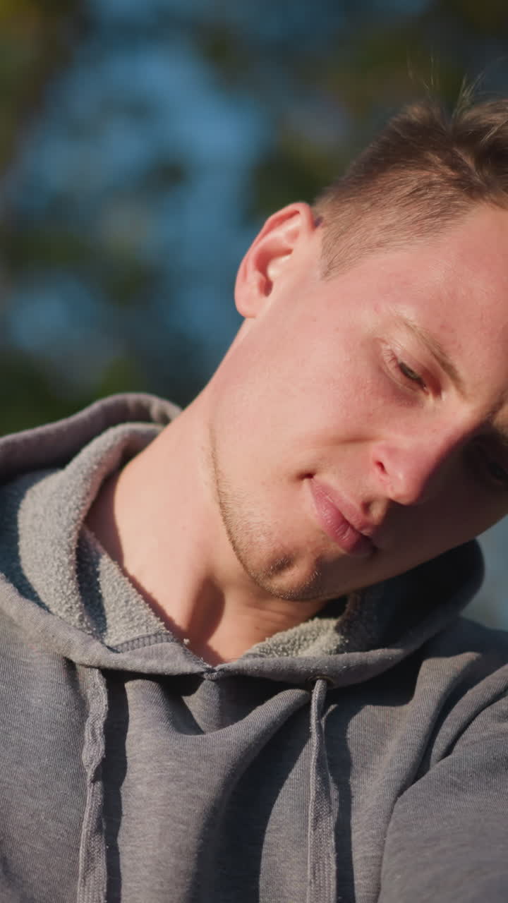 primer plano de un joven con capucha gris, centrado en ajustar los pilotes en un exterior soleado con vegetación borrosa. su expresión concentrada y su cuidadoso agarre del mango del pilote son resaltados por la cálida luz del sol