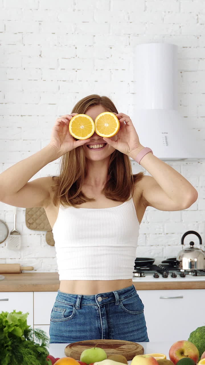 Emotional girl plays with slices of orange. White modern kitchen with fruits and vegetables for a healthy diet. Vertical video