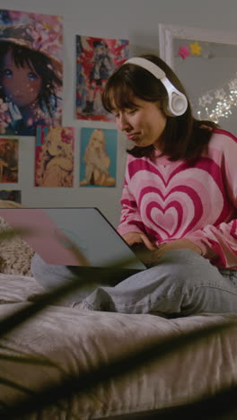 teenage girl listens to music in headphones and scrolls social media using mobile phone lying on the bed in the cozy bedroom. asian girl spending pastime and having fun at home. vertical shot