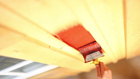 a close-up of a persons's hand painting a wooden plank house wall in red with a paintbrush