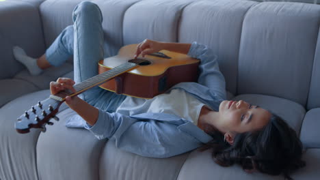 Girl-playing-guitar-on-sofa.-Relaxed-musician-playing-chords-on-acoustic-guitar