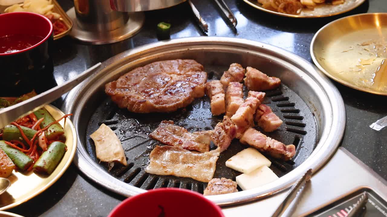 Hand uses tongs to turn pork belly slices on tabletop Korean barbecue grill, daylight setting