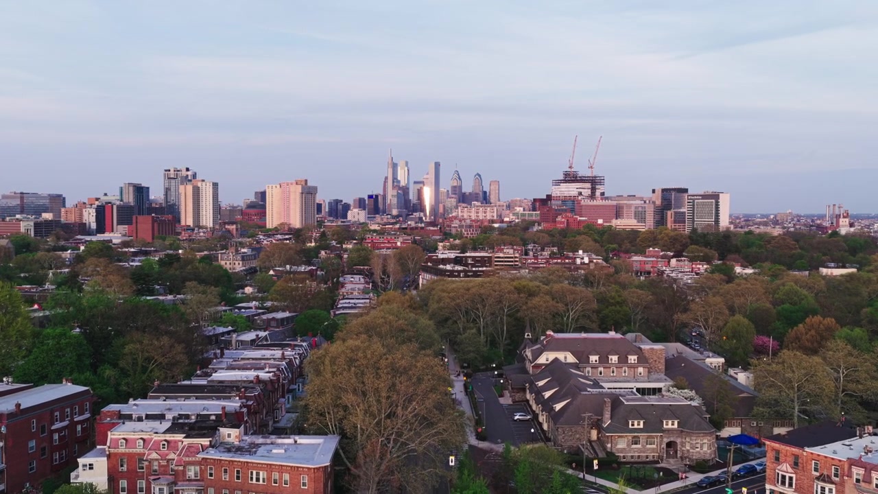 Vista aerea con drone che guarda lungo una strada di Filadelfia verso lo scintillante skyline di Center City al tramonto, con gru edili e quartieri di case a schiera sottostanti