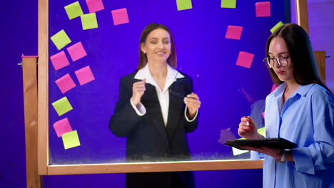 women look at the glass wall talking and discussing stickers. girl at foreground shows the tablet.