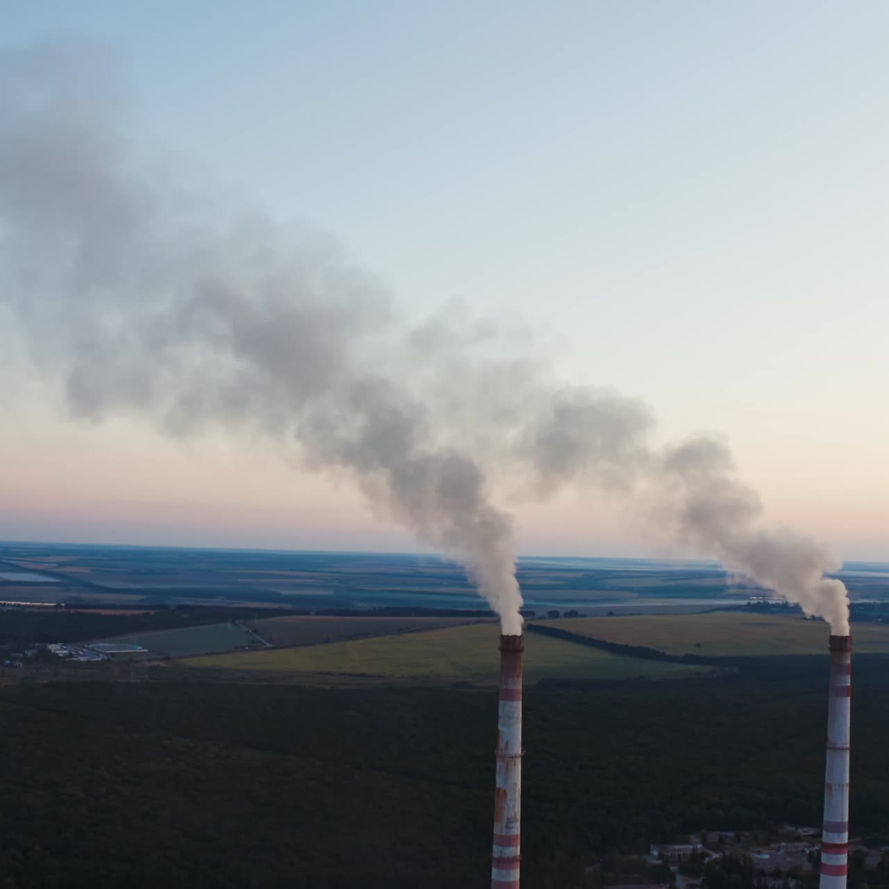 White smoke goes from pipes into the air. Industrial factory produce dirty emissions among the beautiful natural fields in the evening. Aerial view.