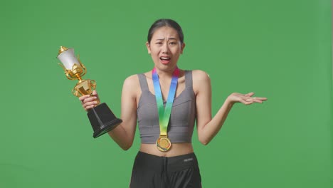 preguntándose mujer asiática con una medalla de oro mirando el trofeo dudosamente en el fondo de la pantalla verde en el estudio