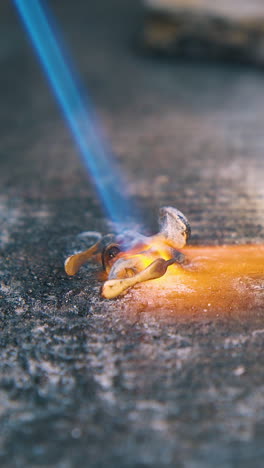 cu: professional jeweler covers gold detail of accessory with material and heats with gas burner on asbestos sheet closeup