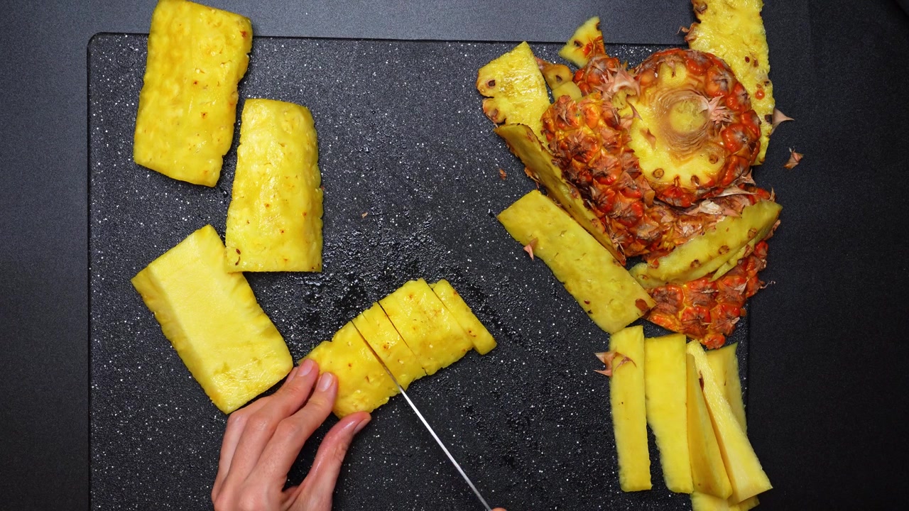 Affettare ananas sbucciato a pezzi su un tagliere. Vista dall'alto di mani che tagliano un ananas sbucciato a fette su un tagliere nero testurizzato.
