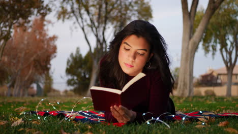 a cute young woman reading a story book laying in the park at twilight in autumn