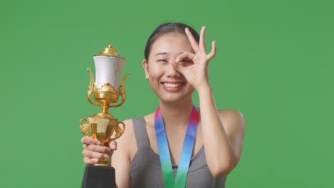 close up of asian woman with a gold medal and trophy showing okay hand sign over eye and smiling to camera as the first winner on green screen background in the studio