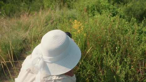 rear view of woman in wide sunhat bending over to photograph and gently touch wild yellow flowers surrounded by tall grass and dense green vegetation under bright morning sunlight