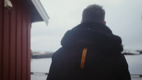 young adult man walk by traditional norwegian red house on coast of lofoten on cold winter day