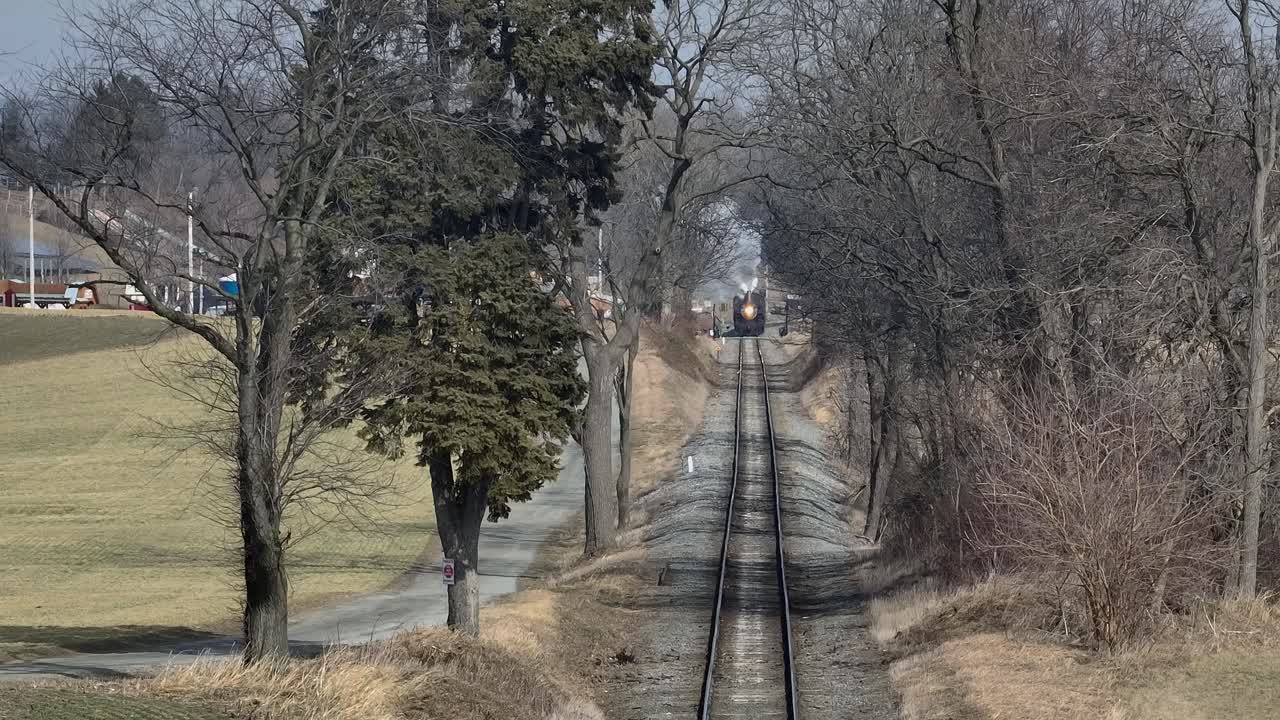 A steam train travels along a set of tracks surrounded by trees in a rural area. The setting features open fields and a quiet road near the railway, creating a serene atmosphere
