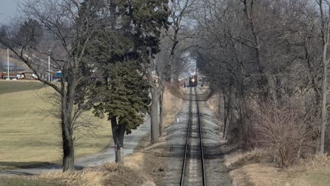 a steam train travels along a set of tracks surrounded by trees in a rural area. the setting features open fields and a quiet road near the railway, creating a serene atmosphere