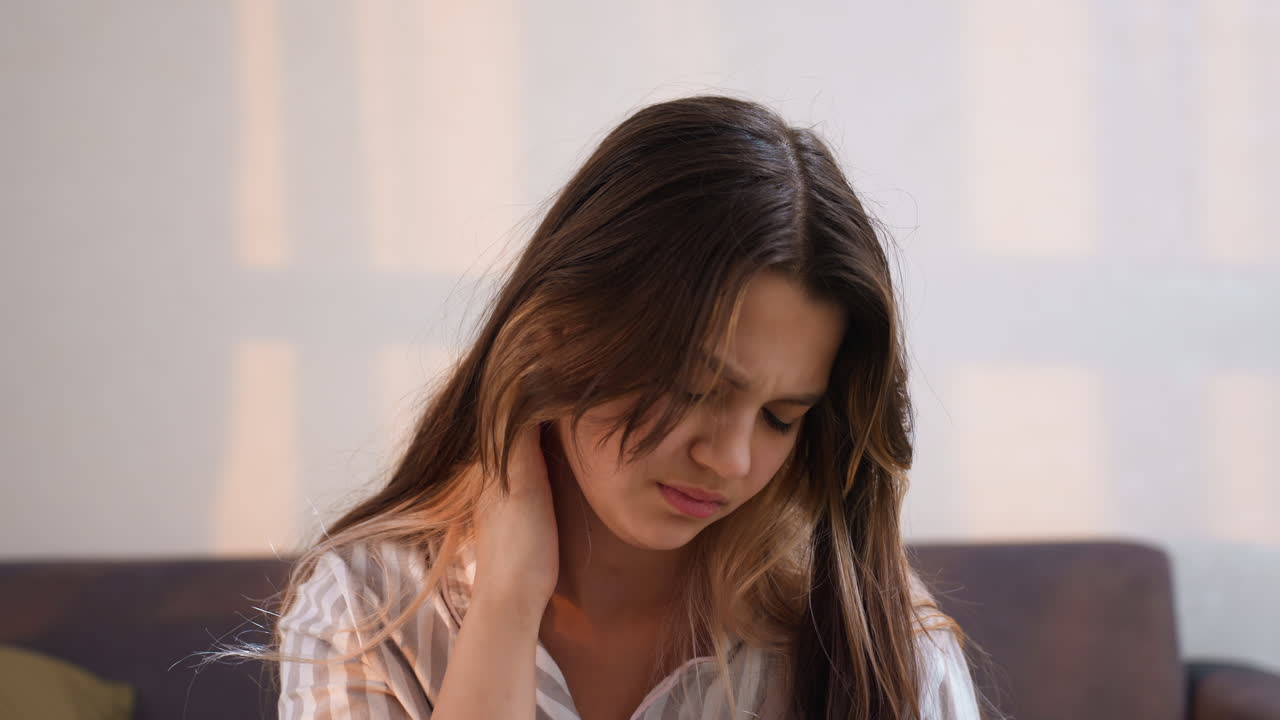 Seated Young Adult Touching Neck On Couch, Warm Late Light Through Window, Casual Shirt And Relaxed Posture, Introspective Expression Alternating With Faint Smile, Quiet Domestic Living Room Scene