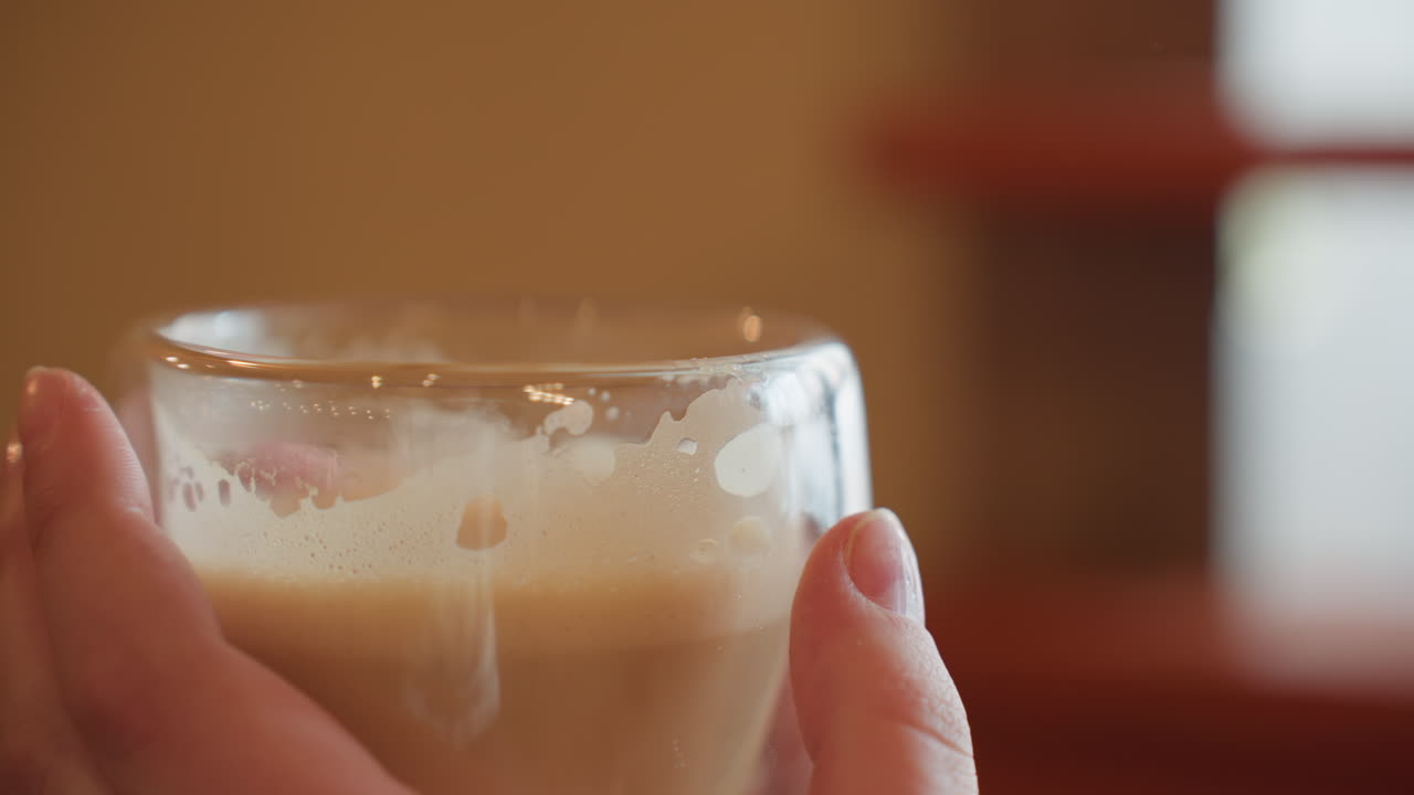 close up of person's hands gently holding transparent glass filled with creamy smoothie indoors, soft light reflecting off surface with warm blurred background