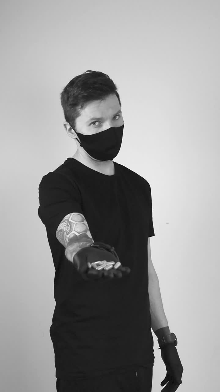 Grayscale footage of a tattooed man with a protective face mask and black rubber gloves showing or offering pills to the viewer while holding the medicine tablets in his palm and moving hand forward.