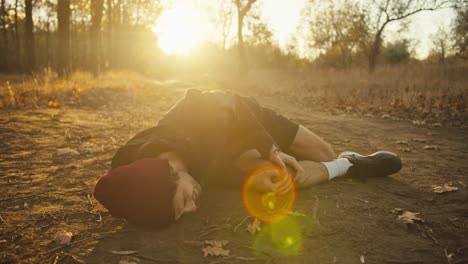 triste hombre cansado atletas con una gorra roja en un uniforme deportivo negro cae al suelo mientras se duele la pierna se aferra a ella y grita mientras corre en el bosque de otoño por la mañana