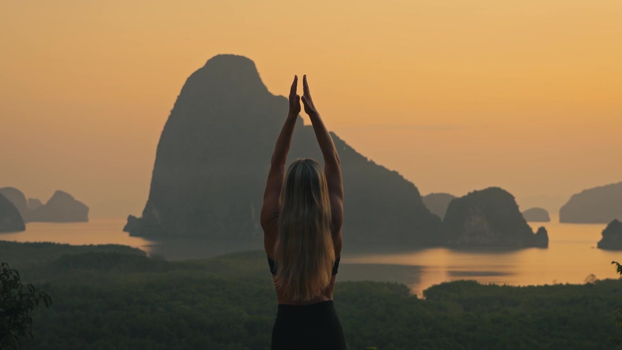 Primo piano di una donna bionda in forma che esegue una posizione di preghiera yoga al punto panoramico di Samet Nangshe, Thailandia. Isole calcaree tropicali e una calda luce dorata creano un'atmosfera di vacanza serena.