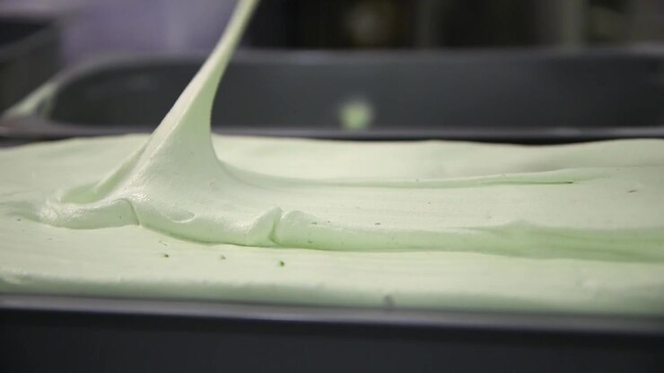 A close-up view of thick, creamy gelato being dispensed from a nozzle into a stainless steel tray, capturing the smooth texture and rich consistency of the dessert.