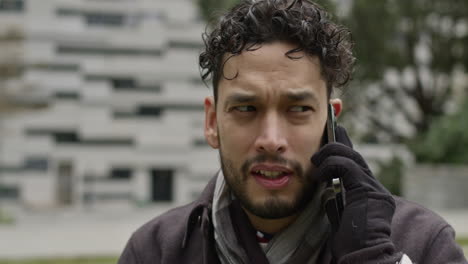 close up of caucasian male model talking with modern new smartphone wearing winter clothes scarf and gloves, background of green park in residential district of smart city, slow motion