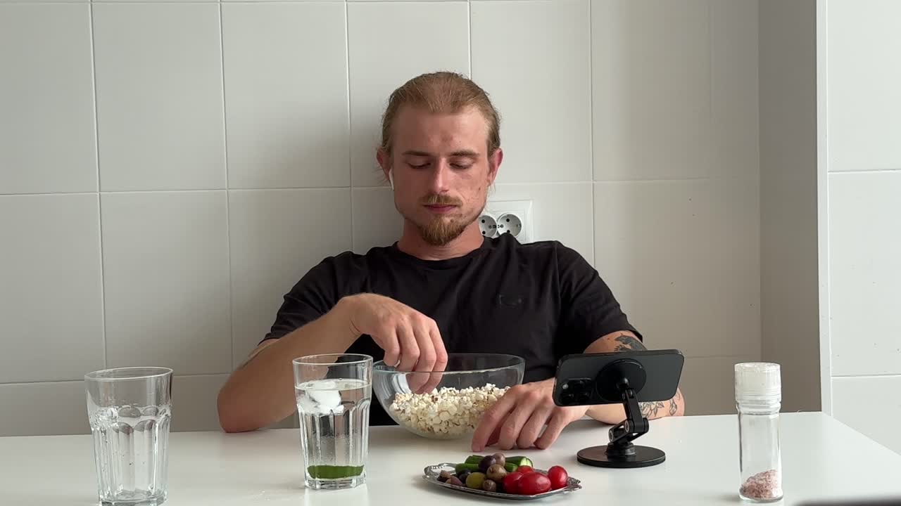 Ung mand spiser popcorn og ser på telefon ved køkkenbordet
