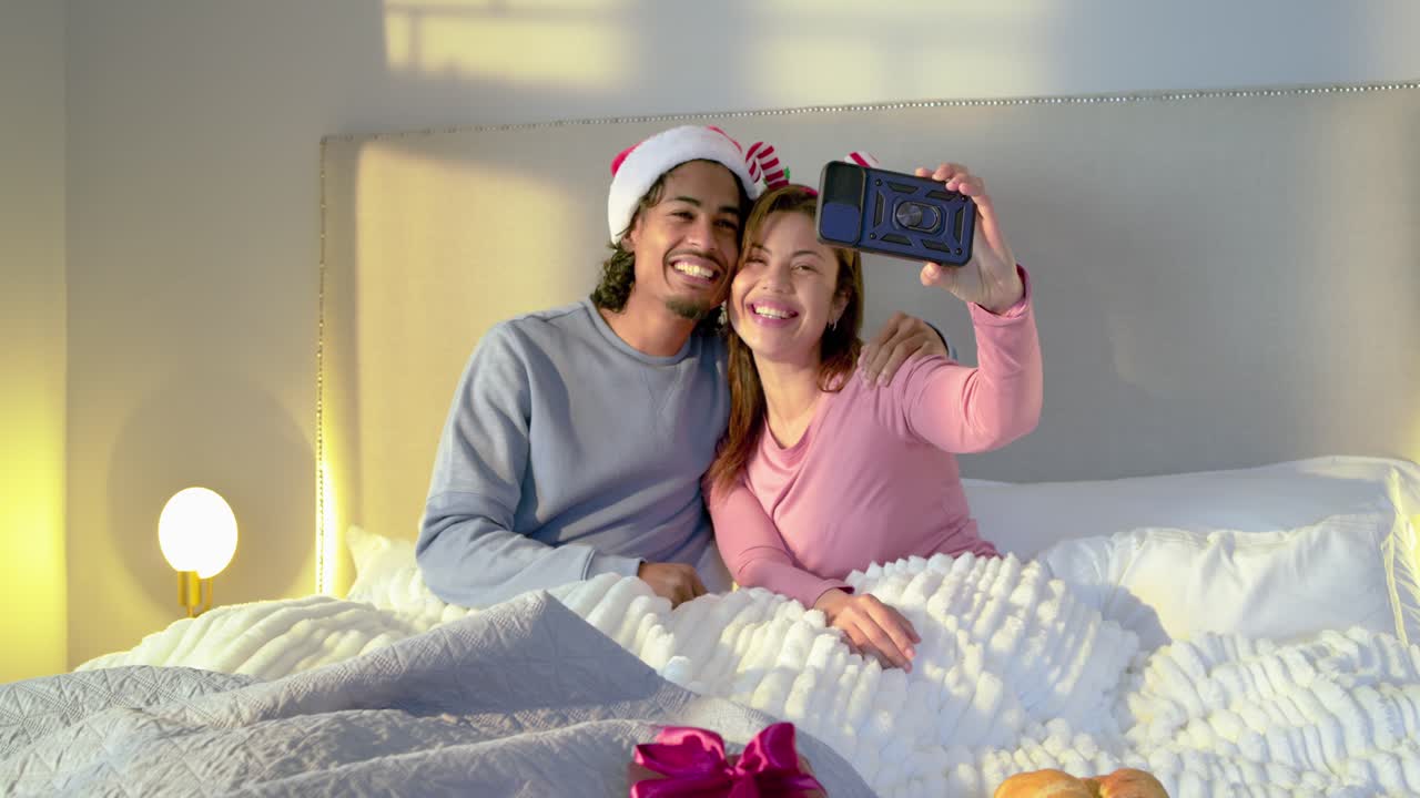 Couple in bed while man in Santa hat placing headband on pink-clad woman snapping smartphone selfie