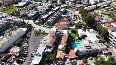 Modern-hotels-with-pools-and-historical-church-of-small-Cretan-town,-aerial-view
