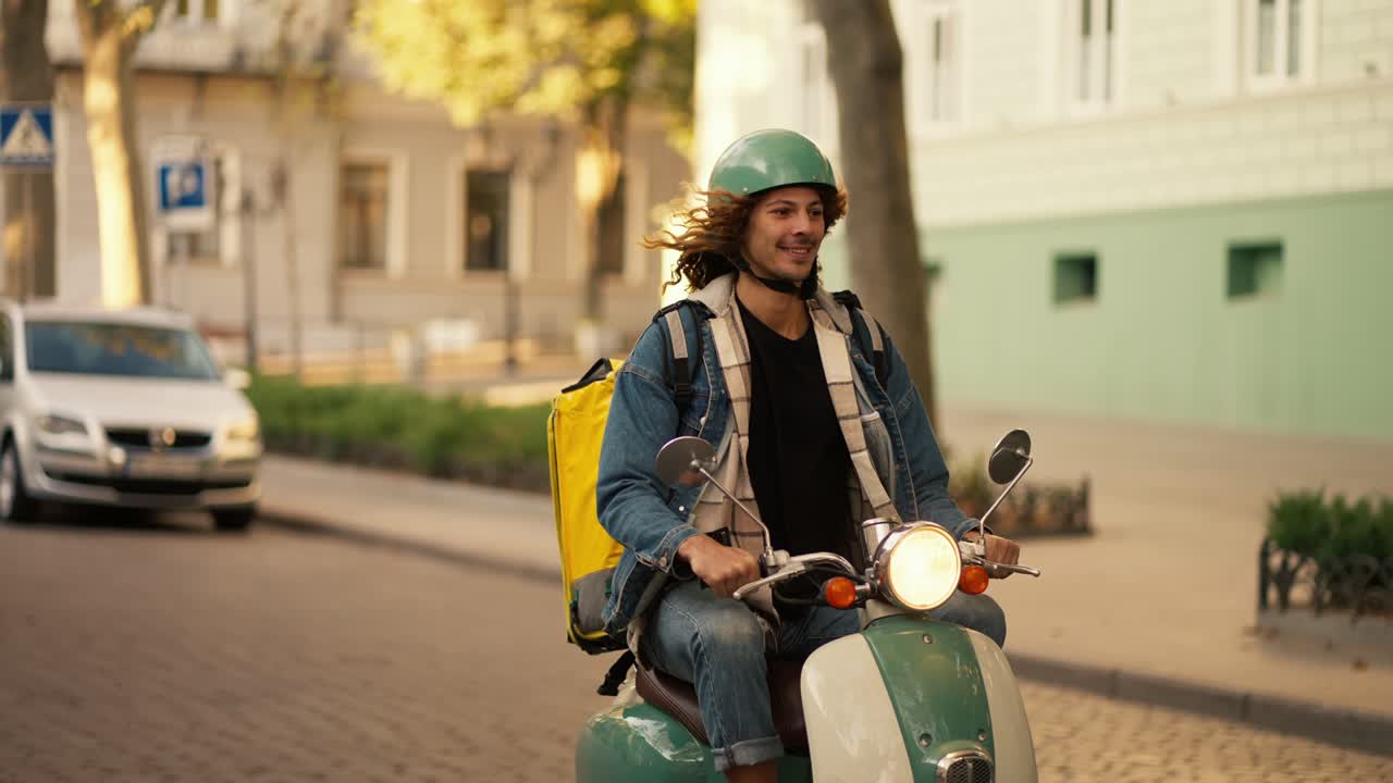 Happy guy with long curly hair in a green helmet and a denim jacket on a green moped with a flashlight on while riding in a summer city