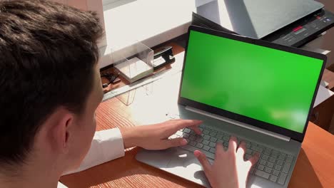un joven está sentado en una computadora portátil con una pantalla verde en la oficina y escribiendo. trabajo, estudio, negocios, finanzas
