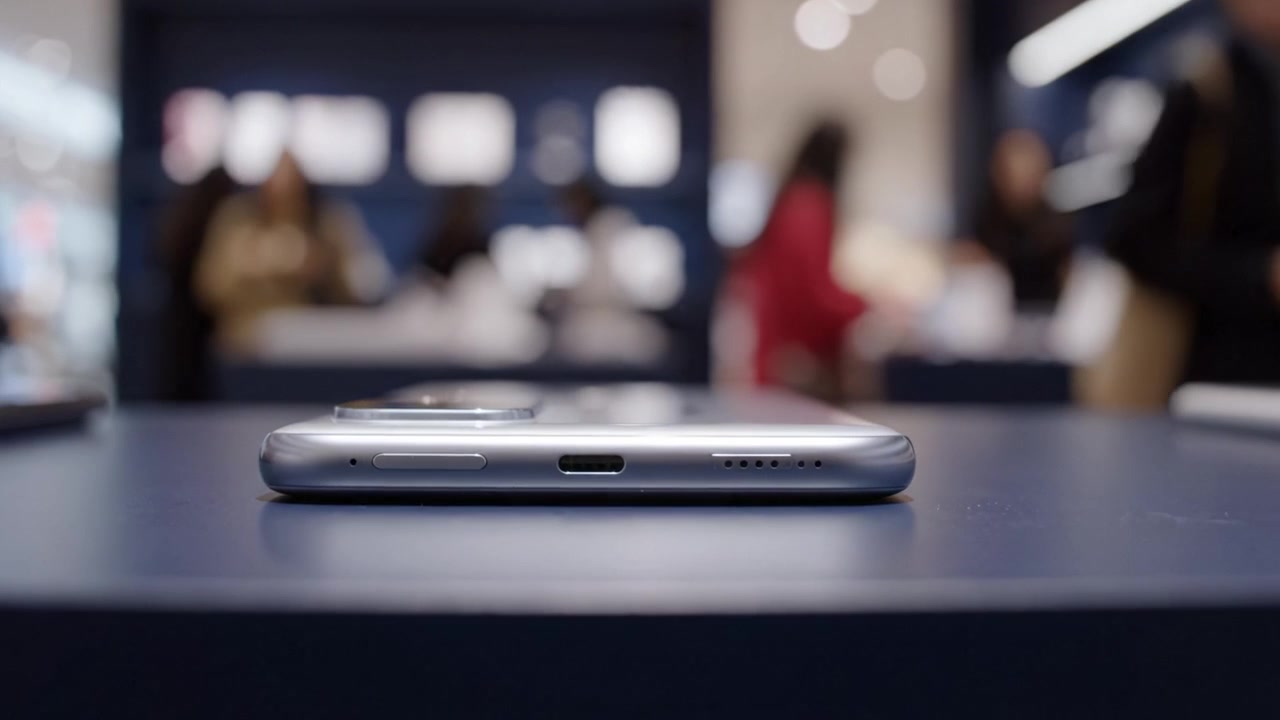 Detailní pohled na chytrý telefon na výstavním stole v maloobchodní prodejně s rozmazanou aktivitou zákazníků v pozadí, představující moderní design mobilního zařízení.