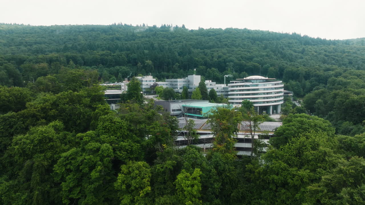 Drone rising backward over DKFZ buildings with solar roofs, forest and hillside backdrop