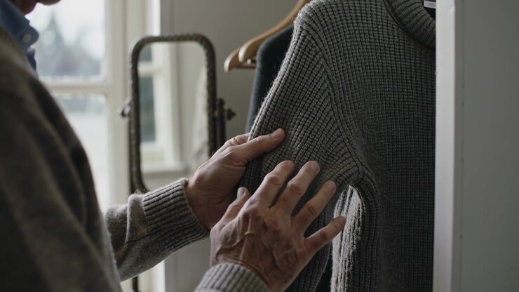 Un uomo anziano con un maglione grigio e un top in maglia leggera ispeziona la cucitura della spalla vicino alla finestra