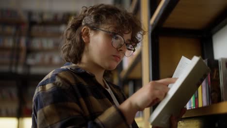 chica segura con cabello rizado que usa gafas lee un libro cerca de los estantes en una biblioteca con muchos libros