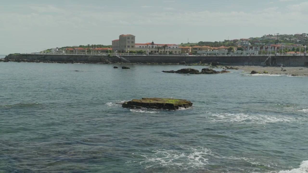 Splendida inquadratura ampia dalla Playa del Rinconín che guarda verso il lungomare di Gijón. Presenta una roccia di scogliera muschiosa isolata nel mare increspato con edifici storici della città e un frangiflutti sullo sfondo.