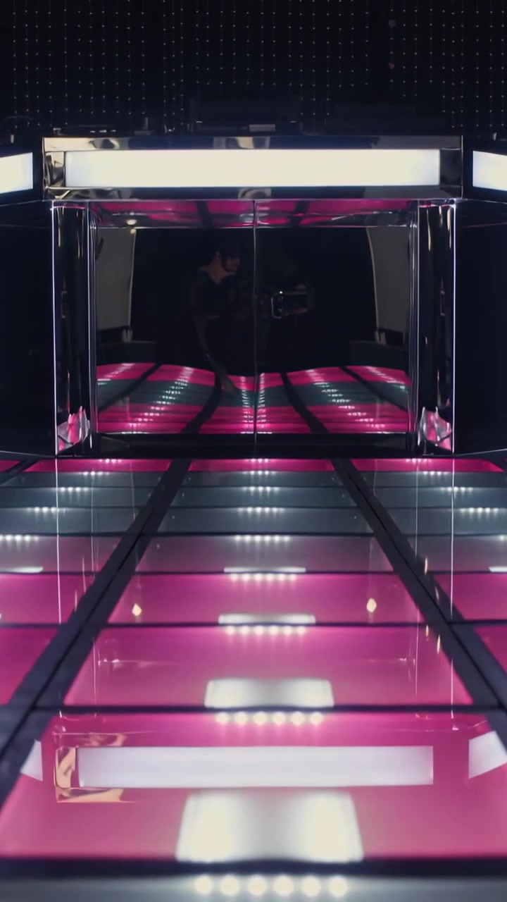 Video verticale: Ripresa in allontanamento che mostra una sala di specchi in una galleria con pavimento illuminato e telefono riflesso.