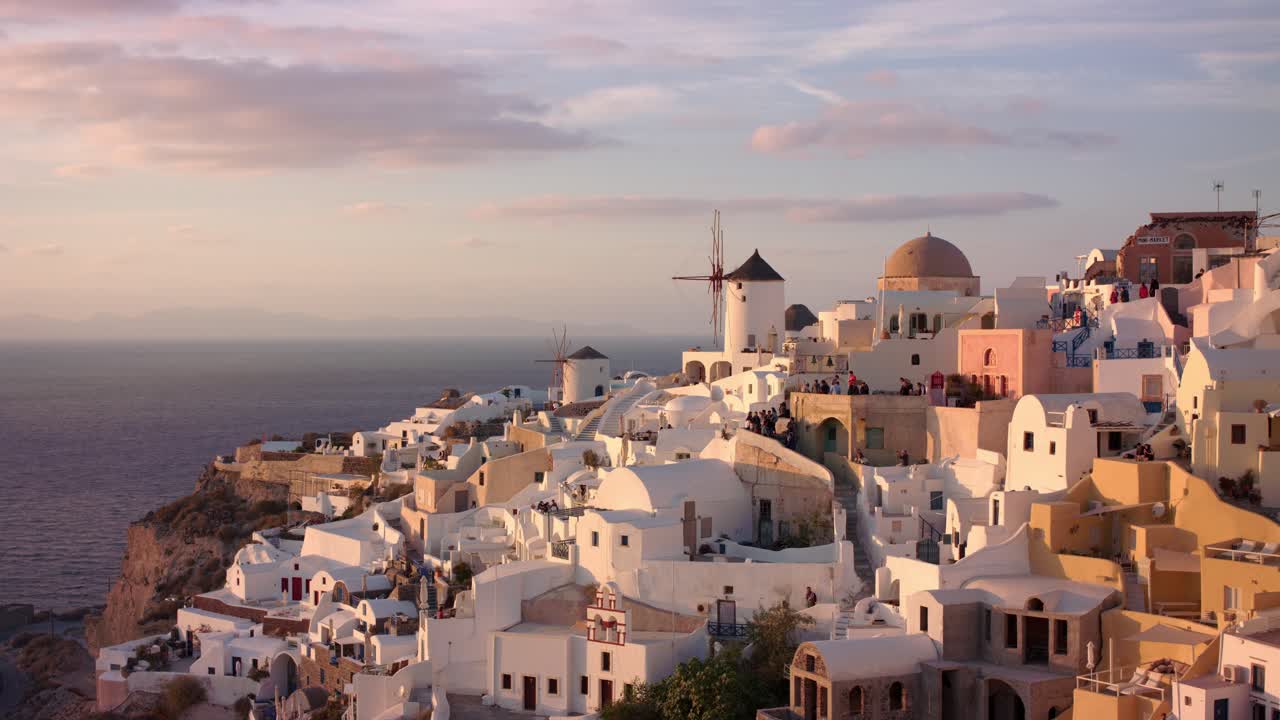 A breathtaking drone view of Santorini’s iconic white buildings and blue domes glowing in golden light at sunset, capturing the island’s unique charm, architecture, and serene Mediterranean beauty